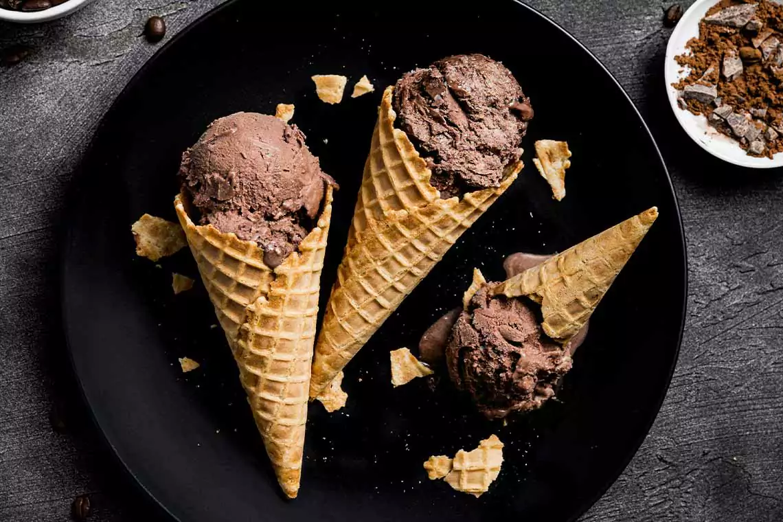 Chocolate ice cream inside a waffle cone on a plate near coffee beans.