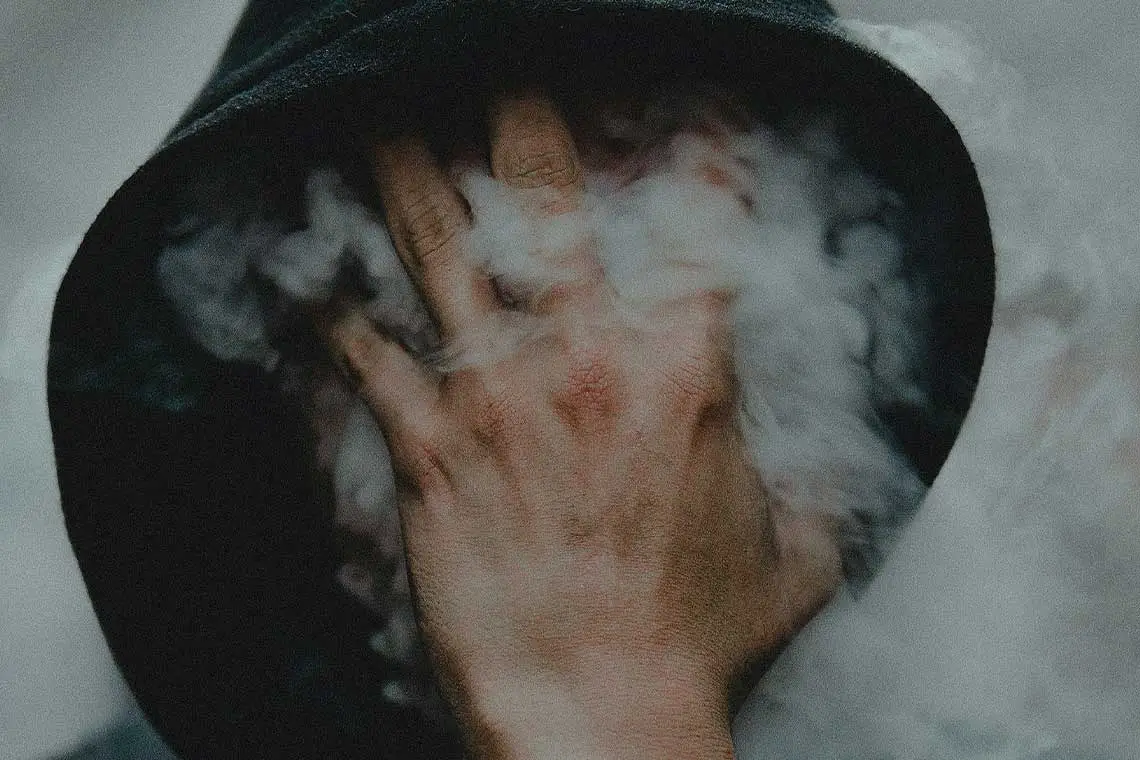 Person with hoodie on covering face with their hand covered in smoke.