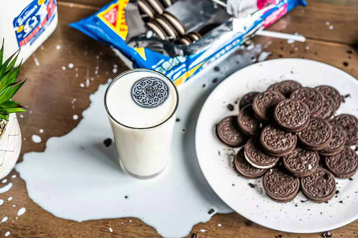 Plate of oreos next to a glass of milk spilling over on to the table.