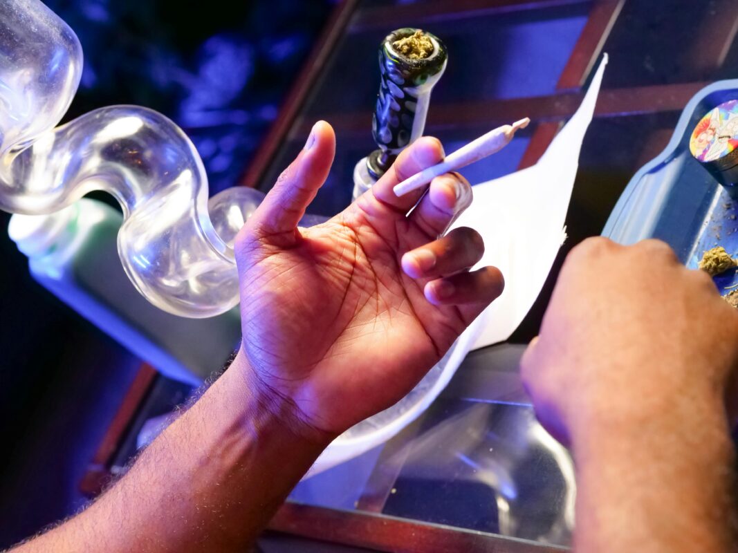 Person holding a joint with a bong and weed in the background